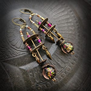 Może przedstawiać: Para ręcznie robionych kolczyków o estetyce vintage. Kolczyki posiadają elementy metalowe w kolorze mosiądzu, w tym okrągłe dyski, koraliki i dekoracyjne akcenty. Każdy kolczyk ma szklaną kulę z kolorowymi, iryzującymi wtrąceniami.