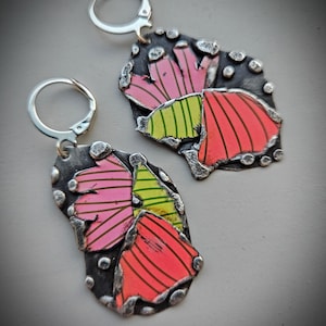 May include: A pair of silver-toned earrings with a floral design. The earrings feature a dark gray, textured base with colorful, striped petals in shades of pink, green, and red. The earrings have a lever-back closure.