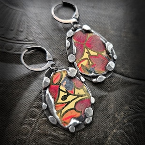 Może przedstawiać: Para srebrnych kolczyków z kwiatowym wzorem. Każdy kolczyk ma kształt łzy z kolorowym wzorem w kolorach czerwonym, żółtym i pomarańczowym. Mają teksturowaną srebrną obwódkę i zapięcie zatrzaskowe.