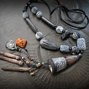 Może przedstawiać: Ręcznie robiony naszyjnik z różnorodnych elementów. Zawiera wzorzyste koraliki, drewniane elementy i centralną zawieszkę. Kolorystyka jest głównie neutralna, z pomarańczowymi i brązowymi akcentami. Unikatowa biżuteria.