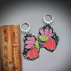 May include: A pair of handmade earrings with a floral design. The earrings are made of silver-toned metal with a dark background and colorful accents. The floral design features pink, red, and green colors. The earrings measure approximately 2 inches in length.