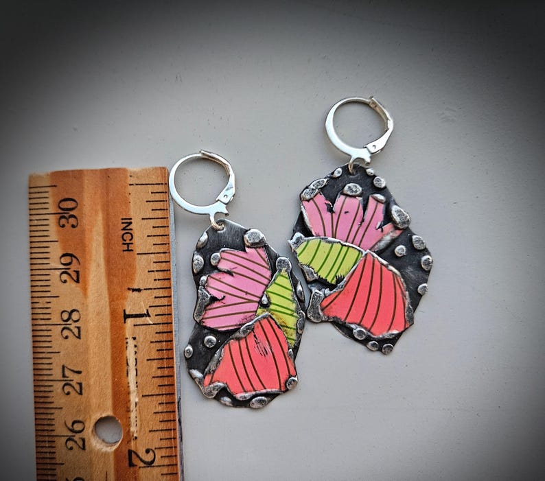May include: A pair of handmade earrings with a floral design. The earrings are made of silver-toned metal with a dark background and colorful accents. The floral design features pink, red, and green colors. The earrings measure approximately 2 inches in length.