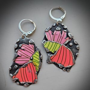 May include: A pair of handmade earrings with a floral design. The earrings feature a dark gray, textured metal base with colorful, abstract flower petals in shades of pink, orange, and green. The earrings have silver-colored lever-back closures.