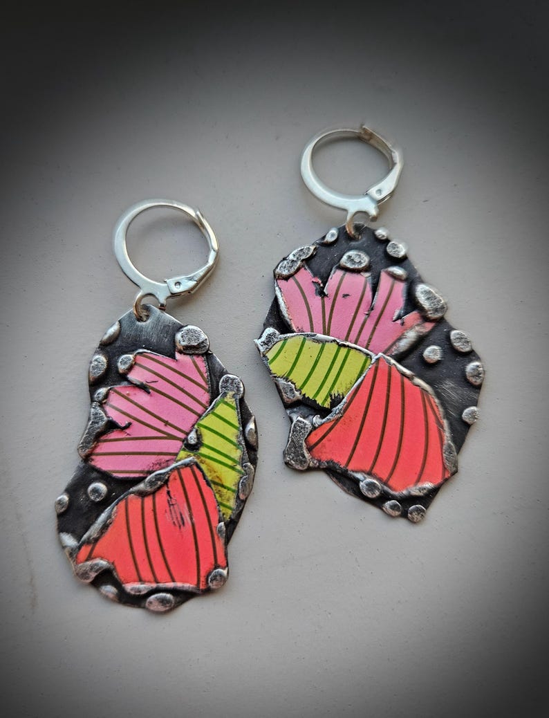 May include: A pair of handmade earrings with a floral design. The earrings feature a dark gray, textured metal base with colorful, abstract flower petals in shades of pink, orange, and green. The earrings have silver-colored lever-back closures.