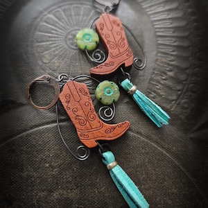 Może przedstawiać: Ręcznie robione kolczyki z brązowymi zawieszkami w kształcie kowbojskich butów z misternymi wzorami. Każdy kolczyk zawiera zieloną koralik kwiatowy, turkusowy frędzel i brązowy haczyk na ucho. Prezentowane na teksturowanej, ciemnej powierzchni.