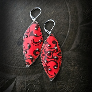 Może przedstawiać: Para czerwonych kolczyków w kształcie łezki z czarnym, wirującym wzorem i srebrnymi ćwiekami. Kolczyki mają srebrne, zawiasowe zapięcia. Biżuteria jest prezentowana na ciemnym tle.