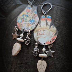 Może przedstawiać: Para unikalnych kolczyków wiszących. Każdy kolczyk ma zawieszkę w kształcie łzy z ilustracją w stylu vintage. Akcenty obejmują gwiazdę, liść i zawieszkę w kształcie szyszki. Kolczyki mają srebrne zapięcia dźwigniowe.