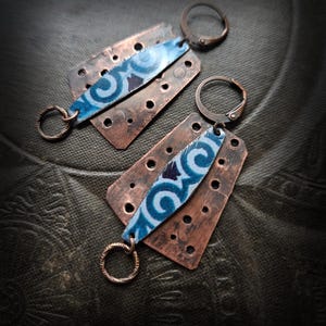 Może przedstawiać: Para ręcznie robionych kolczyków z prostokątami z metalu w kolorze miedzi z małymi otworami. Każdy kolczyk ma niebiesko-biały wzorzysty akcent i okrągłe zapięcie. Kolczyki mają estetykę vintage.