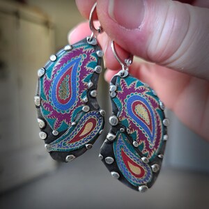 May include: A pair of teardrop-shaped earrings featuring a dark oxidized metal frame with silver-toned studs along the edges. The center displays a vibrant paisley pattern in shades of magenta, teal, blue, and gold, suspended from silver lever-back hooks.