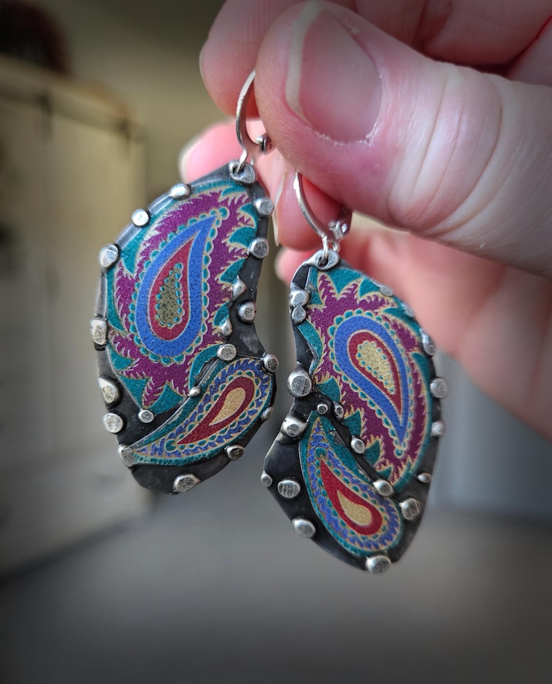 May include: A pair of teardrop-shaped earrings featuring a dark oxidized metal frame with silver-toned studs along the edges. The center displays a vibrant paisley pattern in shades of magenta, teal, blue, and gold, suspended from silver lever-back hooks.