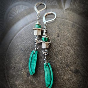 Może przedstawiać: Para srebrnych kolczyków wiszących. Każdy kolczyk ma zielony kamień malachitu, srebrną kostkę i zielono-czarny kamień w paski. Kolczyki mają unikalny, artystyczny design z zapięciami zatrzaskowymi.