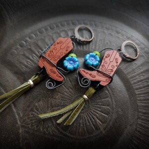 Może przedstawiać: Para ręcznie robionych kolczyków z motywem buta kowbojskiego. Kolczyki wykonane są ze skóry w kolorze brązowym z dekoracyjnymi wirami, niebieskimi akcentami kwiatowymi i oliwkowymi frędzlami. Każdy kolczyk ma srebrny pierścień.