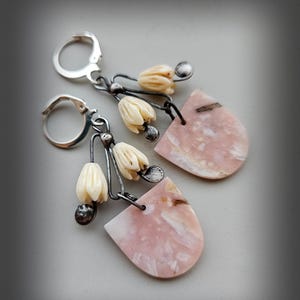 Może przedstawiać: Para kolczyków z wiszącym wzorem z metalu w kolorze srebrnym. Każdy kolczyk ma półokrągły kamień w kolorze bladoróżowym i trzy małe, kremowe zawieszki w kształcie kwiatów. Kolczyki mają zapięcie dźwigniowe.