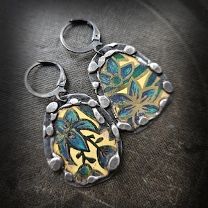 Może przedstawiać: Para ręcznie robionych kolczyków z kwiatowym wzorem. Kolczyki mają srebrną metalową ramkę z teksturowaną obwódką. Środek każdego kolczyka ma złote tło z niebiesko-zielonym kwiatowym wzorem. Kolczyki mają zapięcie zatrzaskowe.