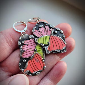 May include: Artistic silver earrings with a colorful floral design. The earrings feature a mix of pink, red, and green hues with black accents. The design includes a lever back closure for secure wear.