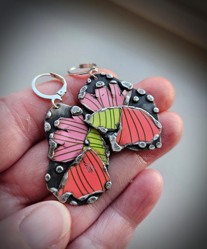 May include: Artistic silver earrings with a colorful floral design. The earrings feature a mix of pink, red, and green hues with black accents. The design includes a lever back closure for secure wear.