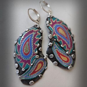 May include: A pair of drop earrings featuring irregular, dark gray metal shapes adorned with colorful paisley patterns in shades of magenta, blue, teal, and yellow. The edges are accented with small, raised silver-toned metal studs.