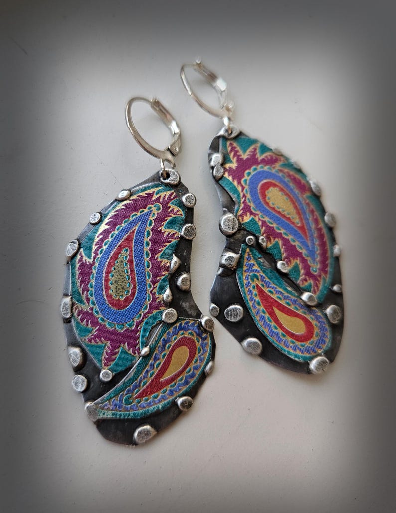 May include: A pair of drop earrings featuring irregular, dark gray metal shapes adorned with colorful paisley patterns in shades of magenta, blue, teal, and yellow. The edges are accented with small, raised silver-toned metal studs.