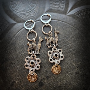 Może przedstawiać: Srebrne kolczyki z zawieszkami w kształcie lamy, kwiatowymi detalami i małymi złotymi monetami. Kolczyki mają wygląd vintage i są prezentowane na ciemnym, teksturowanym tle.