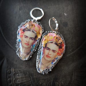 Może przedstawiać: Para srebrnych kolczyków z portretem Fridy Kahlo. Kolczyki mają ozdobną ramkę i zapięcie zatrzaskowe. Portret przedstawia Kahlo z kwiatami we włosach.