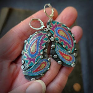 May include: A pair of drop earrings featuring a dark metal base with silver-toned studs along the edges. The surface displays a vibrant paisley pattern in shades of magenta, teal, blue, and gold. Each earring is attached to a silver-toned leverback.