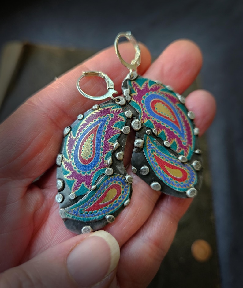 May include: A pair of drop earrings featuring a dark metal base with silver-toned studs along the edges. The surface displays a vibrant paisley pattern in shades of magenta, teal, blue, and gold. Each earring is attached to a silver-toned leverback.