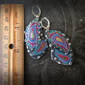 May include: A pair of silver-toned dangle earrings featuring a colorful paisley pattern in shades of magenta, teal, and gold. The earrings are positioned next to a wooden ruler, showing they measure approximately 2.5 inches in total length.