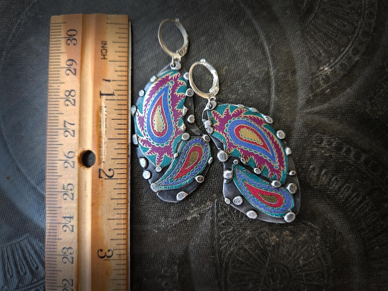 May include: A pair of silver-toned dangle earrings featuring a colorful paisley pattern in shades of magenta, teal, and gold. The earrings are positioned next to a wooden ruler, showing they measure approximately 2.5 inches in total length.