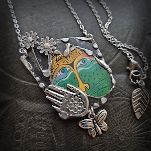 Może przedstawiać: Srebrny naszyjnik z wisiorkiem z kolorowym, artystycznym wzorem twarzy. Wisiorek ma zielone tło z żółtą twarzą, niebieskimi oczami i skomplikowanymi wzorami. Dodatkowe zawieszki to dłoń, motyl i dwa kwiaty przypominające stokrotki.