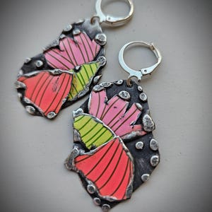May include: A pair of handcrafted earrings with a unique design. The earrings feature a dark, textured metal base with colorful, abstract shapes in pink, red, and green. The earrings have silver lever-back closures. The overall design is artistic and eye-catching.