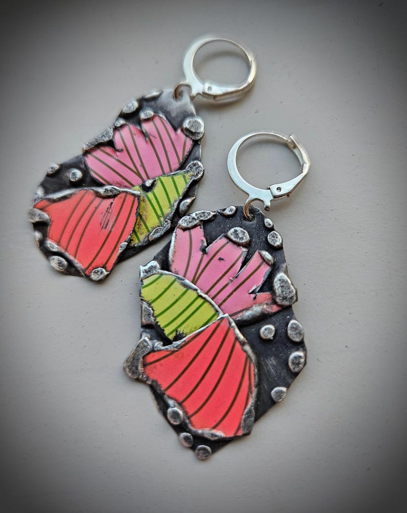 May include: A pair of handcrafted earrings with a unique design. The earrings feature a dark, textured metal base with colorful, abstract shapes in pink, red, and green. The earrings have silver lever-back closures. The overall design is artistic and eye-catching.