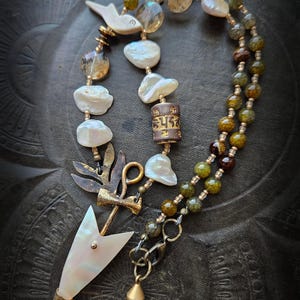 Może przedstawiać: Ręcznie robiony naszyjnik z białymi perłami barokowymi, kamieniami w kolorze bursztynu oraz zielonymi i brązowymi sznurkami z koralików. Naszyjnik posiada ozdobną zawieszkę w kształcie strzały z perłową grotą i złotym stożkowym urokiem.