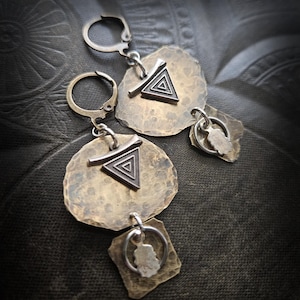 Może przedstawiać: Para ręcznie robionych kolczyków w stylu boho. Każdy kolczyk ma młotkowany, teksturowany, brązowy dysk ze srebrnym trójkątnym wzorem. Poniżej wisi mniejszy, srebrny urok z brązowego kwadratu.