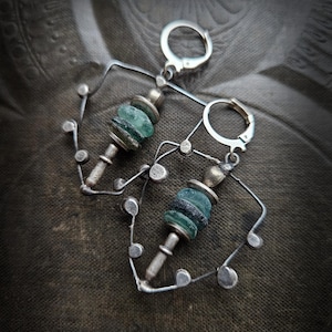 Może przedstawiać: Para ręcznie robionych srebrnych kolczyków. Każdy kolczyk ma centralną kolumnę z ułożonych, nieregularnych, turkusowych szklanych koralików. Koraliki są utrzymywane przez srebrne elementy metalowe. Kolczyki mają unikalny, geometryczny design.