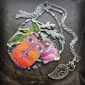 Może przedstawiać: Ręcznie robiony naszyjnik z wisiorkiem z kolorowym, artystycznym wzorem twarzy. Wisiorek ma srebrną metalową ramkę i pasujący łańcuszek. Projekt twarzy obejmuje odcienie pomarańczu, różu i błękitu, z akcentami liści i kwiatów.