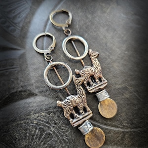 Może przedstawiać: Ręcznie robione kolczyki z detalicznym wzorem lamy w odcieniach brązu i srebra. Kolczyki mają okrągły kształt i mały, okrągły, drewnopodobny akcent na dole. Na ciemnym, teksturowanym tle.