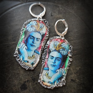 Może przedstawiać: Para srebrnych kolczyków z portretem Fridy Kahlo. Kolczyki mają prostokątny kształt z ozdobną obwódką. Portret jest w odcieniach niebieskiego, czerwonego i żółtego, z zapięciem zatrzaskowym.