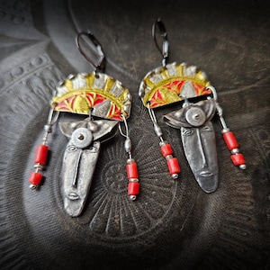 Może przedstawiać: Para ręcznie robionych kolczyków z motywem maski plemiennej. Kolczyki mają twarze z metalu w odcieniach srebra z czerwonymi i żółtymi akcentami oraz czerwonymi koralikami. Unikalny i artystyczny design.