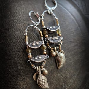 Może przedstawiać: Ręcznie robione srebrne kolczyki o unikalnym designie. Każdy kolczyk ma trzy srebrne elementy w kształcie liści, zaakcentowane małymi zielonymi i złotymi koralikami. Małe zawieszki i koraliki zwisają z dołu, tworząc warstwowy, bohemiczny wygląd.