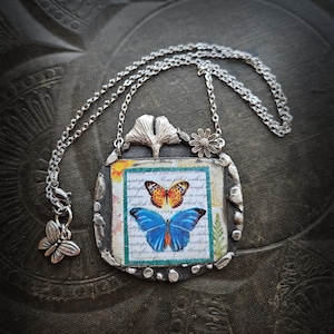Może przedstawiać: Srebrny naszyjnik z wisiorkiem, prostokątny wisiorek z motywem motyla. Wisiorek ma srebrną ramkę i łańcuszek z zawieszką w kształcie motyla. Motyw motyla zawiera pomarańczowego i niebieskiego motyla.