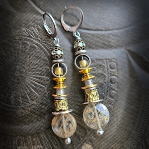 Może przedstawiać: Kolczyki wiszące o warstwowym designie. Kolczyki posiadają srebrne obręcze i kombinację złotych i srebrnych koralików, z przezroczystymi, okrągłymi, teksturowanymi koralikami na dole. Kolczyki mają estetykę vintage lub boho.