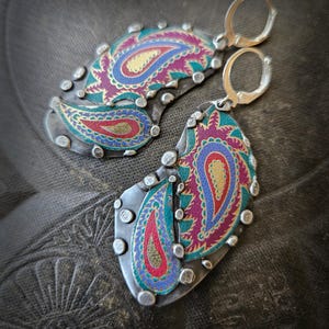 May include: A pair of dangle earrings featuring oxidized silver metal bases with raised silver dots along the edges. The center of each earring displays a colorful paisley pattern in shades of magenta, teal, blue, and gold with a textured finish.