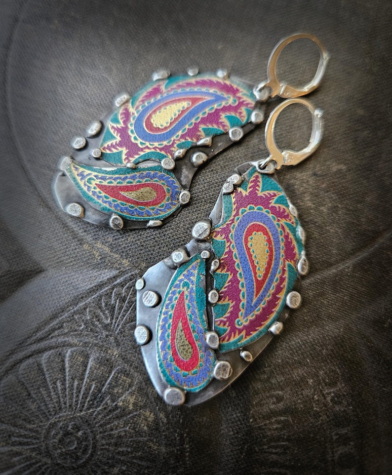 May include: A pair of dangle earrings featuring oxidized silver metal bases with raised silver dots along the edges. The center of each earring displays a colorful paisley pattern in shades of magenta, teal, blue, and gold with a textured finish.