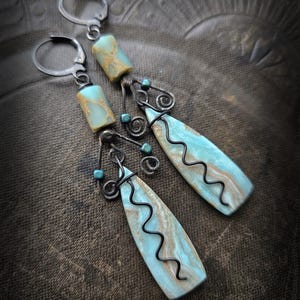 Może przedstawiać: Para ręcznie robionych kolczyków z turkusowymi i brązowymi prostokątnymi i łezkowatymi kamieniami. Kolczyki mają srebrne haczyki, czarne druciane akcenty i małe niebieskie koraliki. Kamienie w kształcie łezki mają czarny falisty wzór linii.