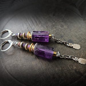 Może przedstawiać: Para kolczyków z prostokątnymi, przezroczystymi fioletowymi kamieniami szlachetnymi. Kolczyki mają srebrne zapięcia typu bigiel, złote i srebrne akcenty oraz detale łańcuszka. Kamienie mają około 1 cm długości.