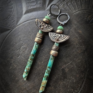 Może przedstawiać: Para ręcznie robionych kolczyków z prostokątnymi kamieniami w kolorze turkusowym. Kolczyki mają srebrne metalowe akcenty, w tym dekoracyjne półksiężyce i małe zielone koraliki. Kolczyki są prezentowane na ciemnej, teksturowanej powierzchni.