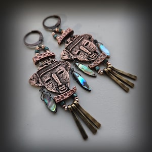 Może przedstawiać: Miedziane kolczyki z motywem stylizowanej twarzy i misternymi detalami. Każdy kolczyk ma iryzujące akcenty i mosiężne pręciki. Kolczyki mają zapięcie na haczyk. Długość ok. 7 cm.