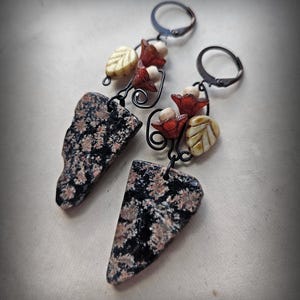 Może przedstawiać: Para ręcznie robionych kolczyków z czarno-brązowymi, nakrapianymi kamiennymi zawieszkami. Kolczyki mają elementy dekoracyjne, w tym czerwone koraliki w kształcie kwiatów, kremowe koraliki w kształcie liści i czarne druciane akcenty. Zapięcie na zatrzask.