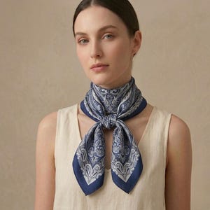 Peut inclure: Un foulard en soie bleu marine et blanc avec un motif cachemire noué autour du cou. Le foulard présente un motif détaillé avec une bordure bleu foncé. Le foulard est porté sur un haut crème.