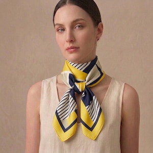 Peut inclure: Un foulard en soie jaune, marine et blanc, avec un motif géométrique. Le foulard est noué au cou et présente une combinaison de rayures et de blocs de couleur unis. Le foulard est porté sur un haut crème.